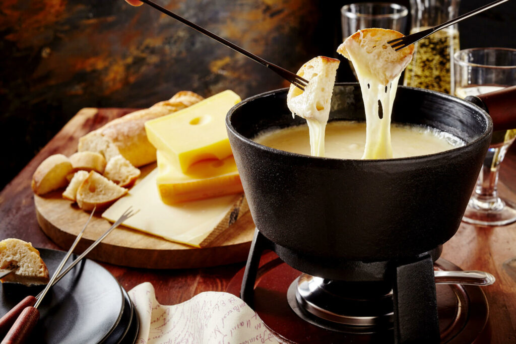 Caquelon de fondue avec deux morceaux de pain accroché à du fromage fondant ; avec un plateau de fromages à l'arrière plan